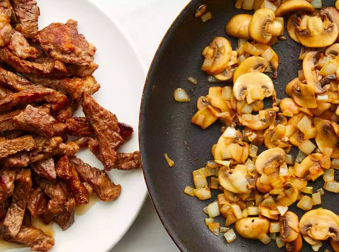 Aerial view of mushrooms and onions cooking in a skillet next to a plate of cooked beef.