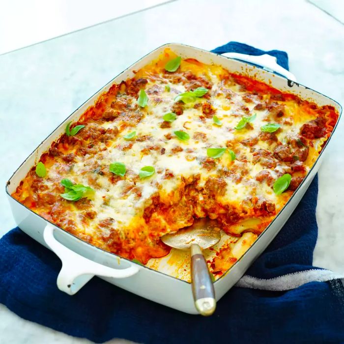 Close-up of lasagna with ricotta bechamel, garnished with fresh basil, in a white baking dish, placed on a navy kitchen towel.