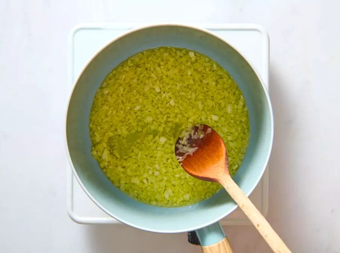 A pan with chopped onion and garlic sizzling in oil, being stirred with a wooden spoon