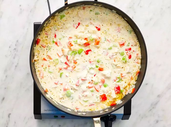 A top-down view of sour cream being added to a skillet filled with onions, chicken, red bell pepper, celery, and chicken stock