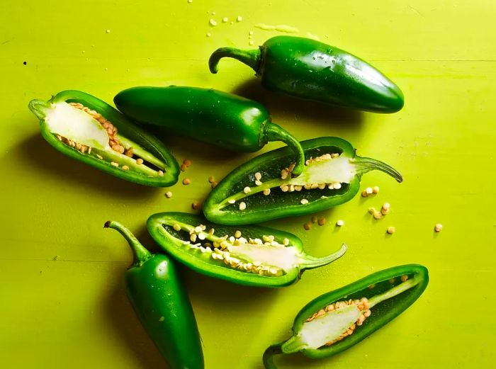 A collection of jalapeño peppers on a vibrant green backdrop, with some sliced open to reveal their seeds.