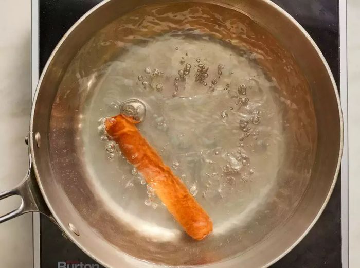 A bird's-eye view of a single hot dog simmering in a pot of water.