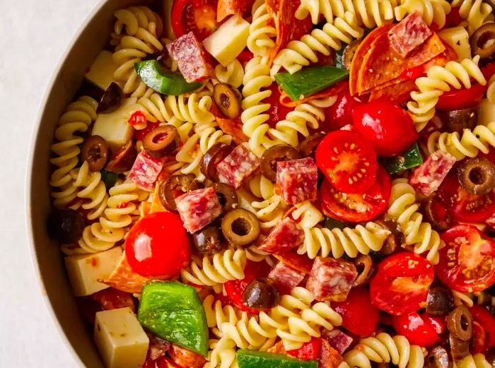 close-up of a large bowl filled with pasta salad, including pasta, tomatoes, salami, pepperoni, peppers, and cheese