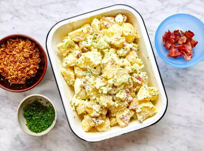 A bowl of Million Dollar Potato Salad, served with chopped chives, crispy bacon, and golden fried onions on the side