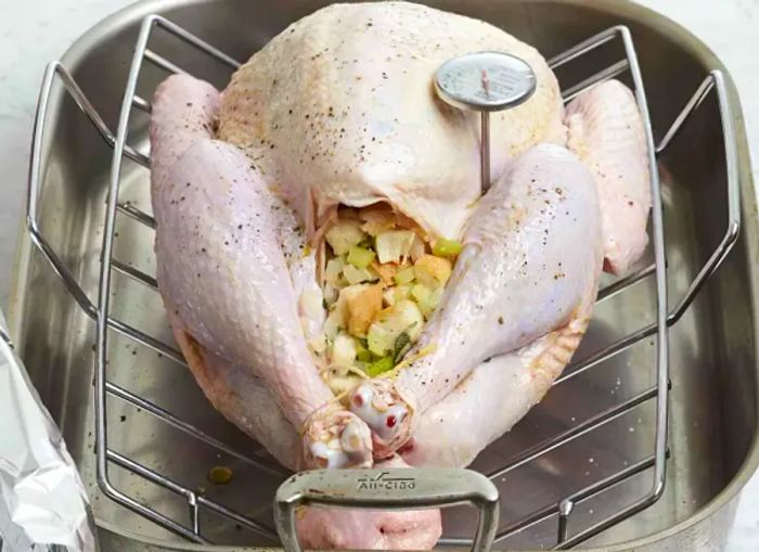 An unroasted stuffed turkey resting on a rack in a roasting pan with a meat thermometer inserted into the thigh