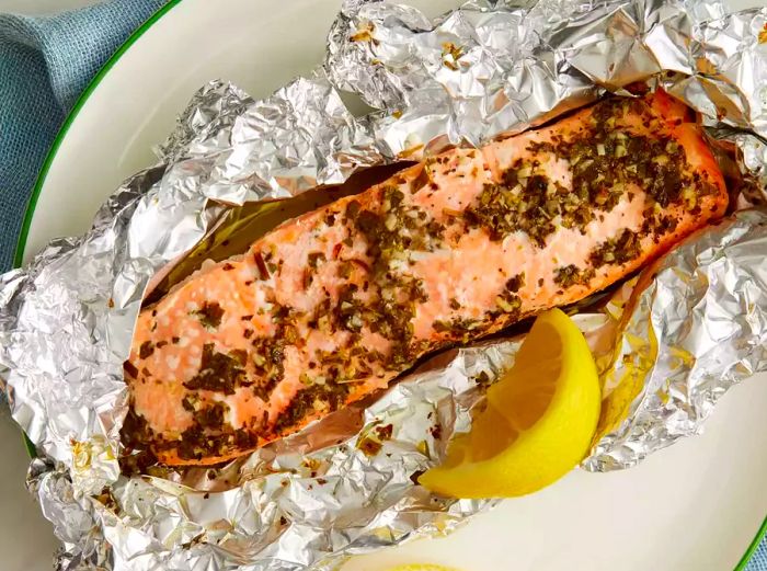 An overhead view of a baked salmon fillet wrapped in tin foil, accompanied by a side of lemon wedges.