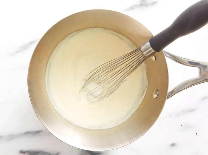 An overhead shot of a pan with milk being whisked into a butter and flour roux to create a creamy béchamel sauce.