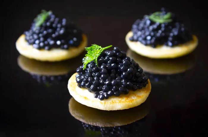 Three blinis topped with black caviar and fresh dill, set against a dark background.