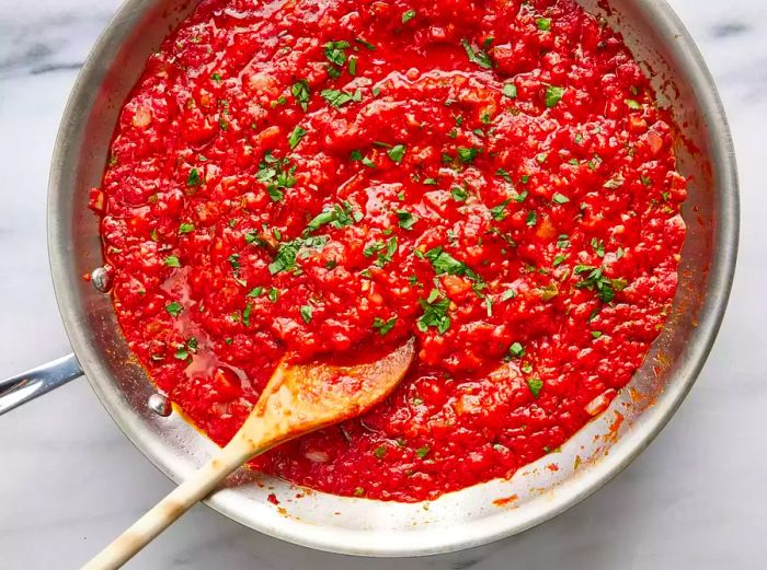 A close-up of marinara sauce with fresh herbs and a wooden spoon in a skillet