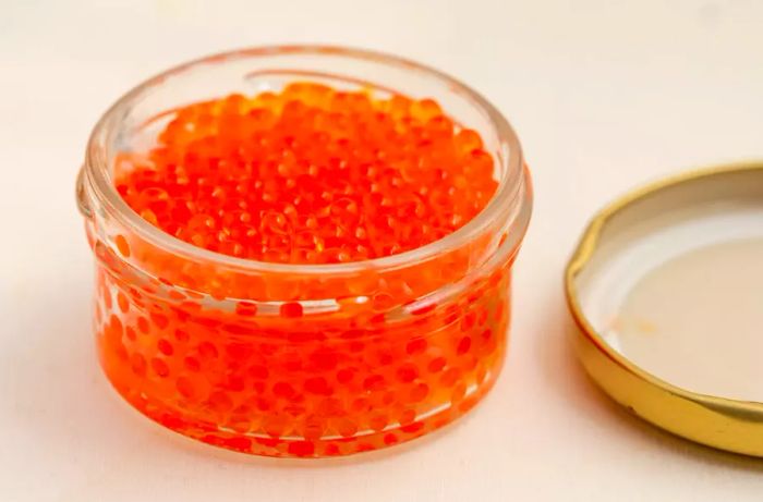 A partially opened jar of red caviar made from trout roe, with the lid slightly visible, set on a wooden poplar surface.