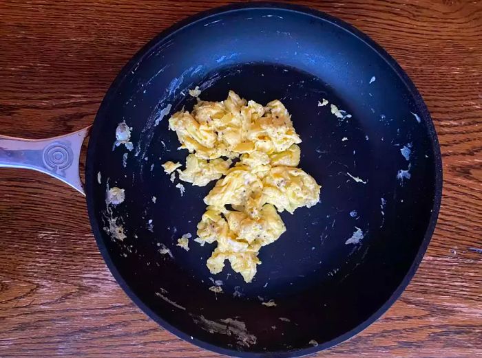 A pile of scrambled eggs rests in the center of a nonstick skillet, with some leftover egg residue stuck to the pan.