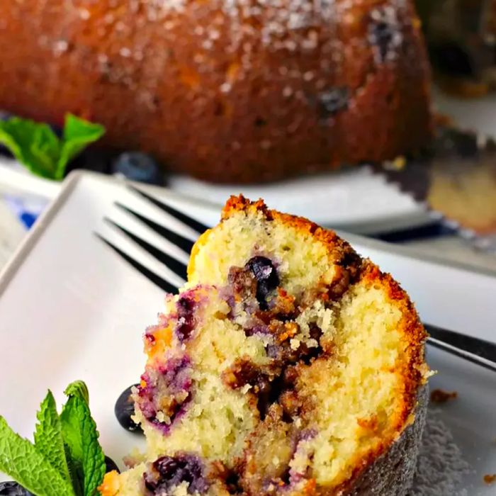 Close-up of a slice of Blueberry Sour Cream Coffee Cake on a white plate with a fork, with another whole Blueberry Sour Cream Coffee Cake in the background