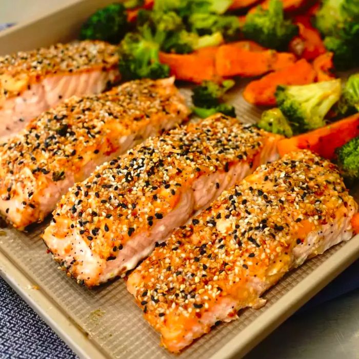 Four salmon fillets seasoned with 'everything bagel' seasoning, paired with roasted broccoli florets and sweet potato wedges on a sheet pan.