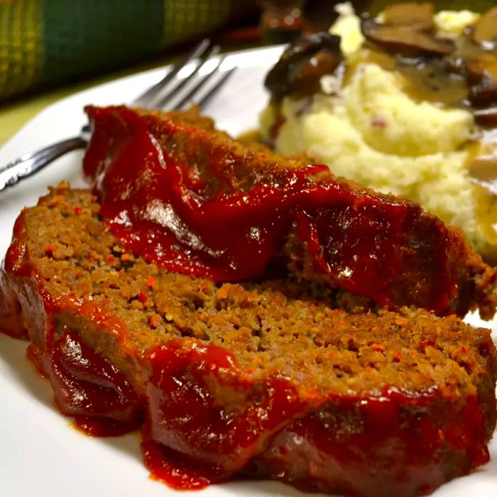 Two slices of ketchup-glazed meatloaf served on a white plate, with mashed potatoes in the background