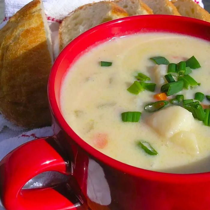 A red bowl filled with creamy soup, loaded with chunks of potato and topped with sliced green onions, with a side of sliced bread.