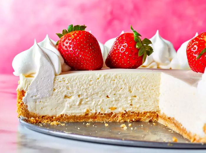 A close-up of a no-bake cheesecake sitting on a pan, with a quarter of it missing, topped with cream and fresh strawberries.