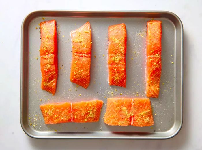 A baking sheet filled with salmon fillets, seasoned with lemon pepper, garlic powder, and salt.