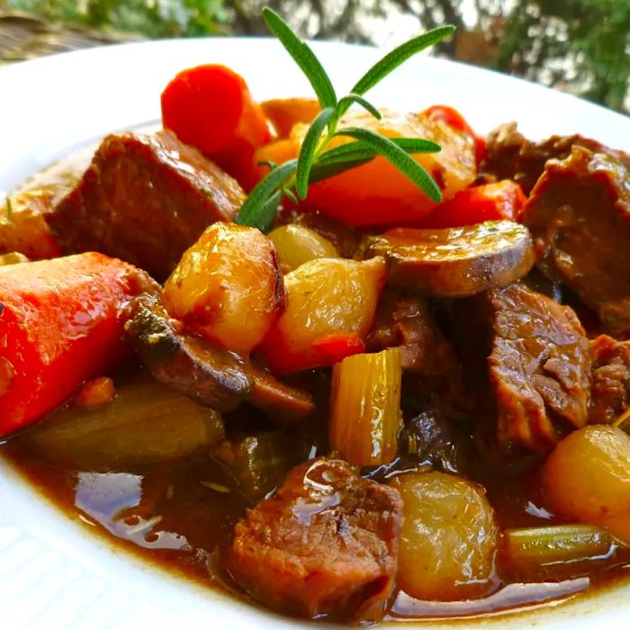 Close-up of a white bowl filled with chunks of beef, pearl onions, carrots, and potatoes, garnished with a sprig of rosemary