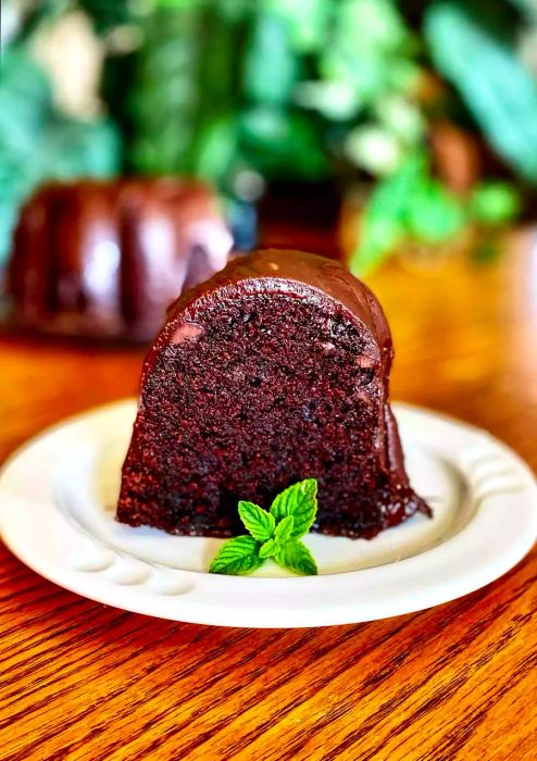 A slice of rich dark chocolate Bundt cake on a white plate, garnished with a sprig of mint