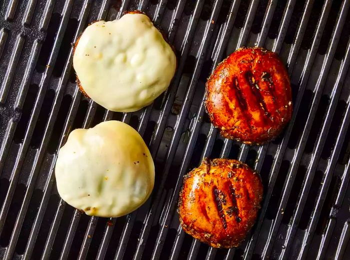 Four grilled portobello mushroom caps on the grill grates, two of which are topped with melted cheese.