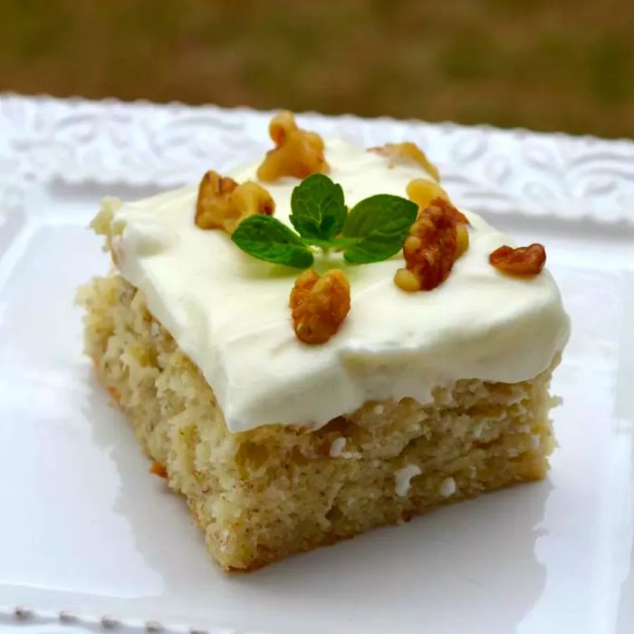 A square slice of banana cake topped with smooth cream cheese frosting, garnished with chopped walnuts and a sprig of fresh mint