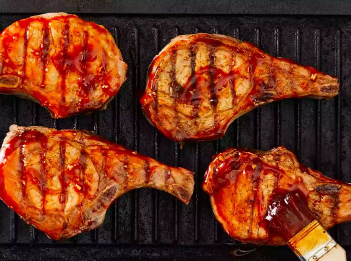 An overhead shot of four pork chops on a griddle, with sauce being brushed onto one of the chops.