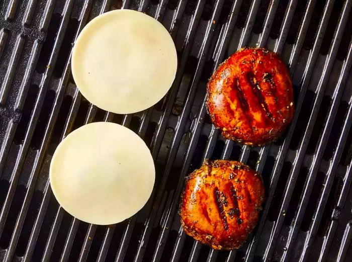 Four grilled and marinated portobello mushroom caps on the grill grates, with two topped with slices of cheese.