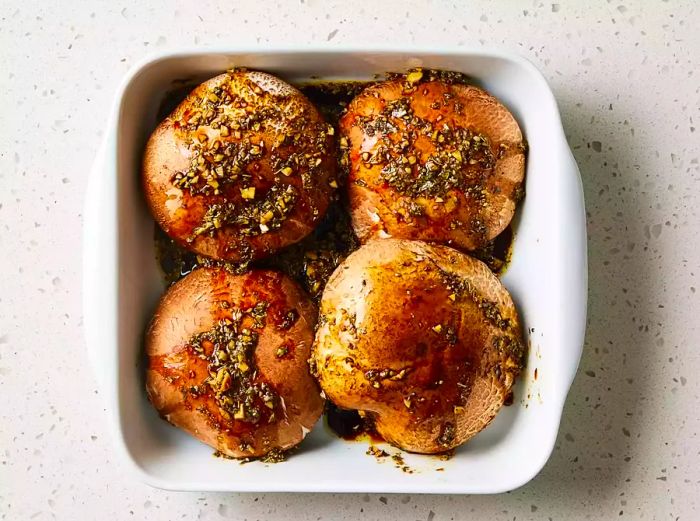 A shallow dish containing four portobello mushroom caps, drizzled with vinaigrette.