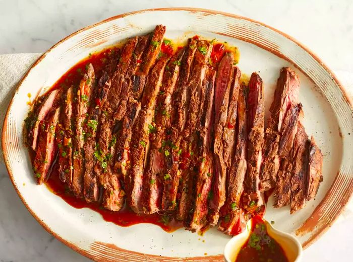 A top-down view of grilled, marinated steak, sliced and served on a platter with the reserved marinade poured over the top.