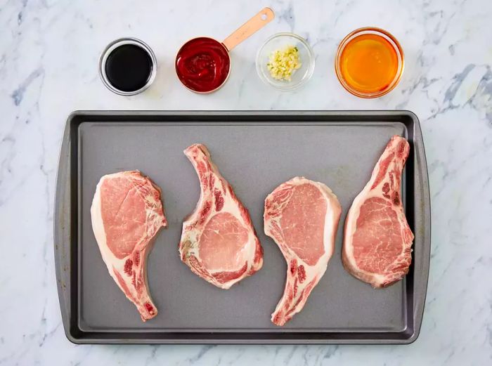 Four raw, bone-in pork chops placed on a baking sheet, accompanied by four small bowls containing the ingredients.