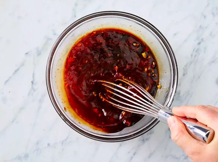 An overhead shot of brown sauce in a clear glass bowl with a hand using a whisk to stir the sauce.