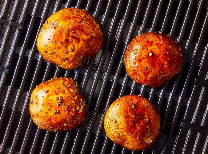 Four marinated portobello mushroom caps sizzling on the grill grates.