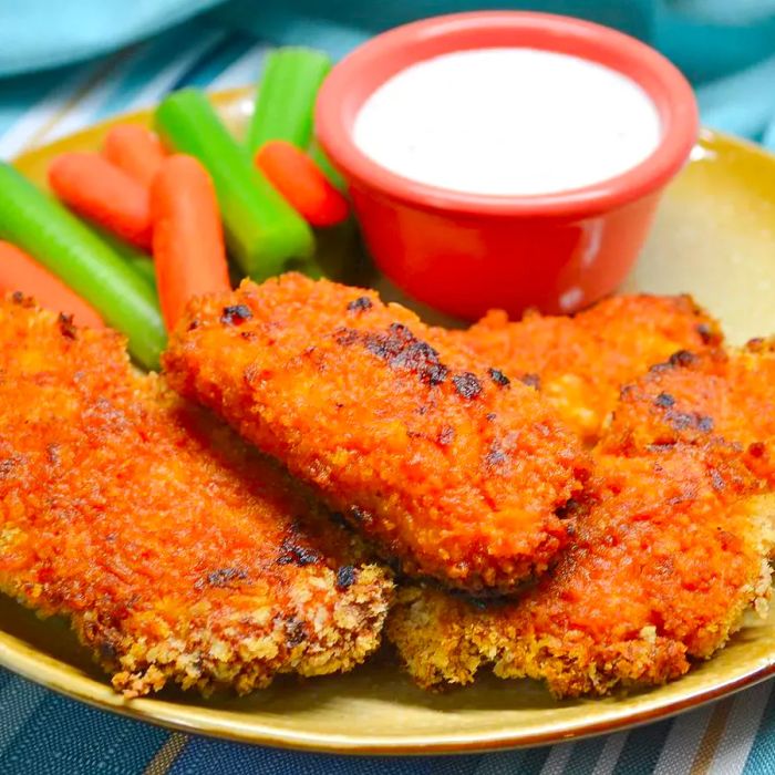 A close-up of crispy Buffalo Chicken Strips served alongside carrots, celery, and a dipping sauce in a red cup.