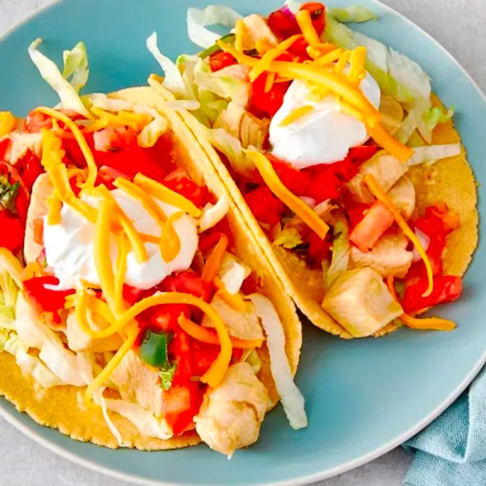 Two chicken tacos on corn tortillas, topped with onions, tomatoes, sour cream, and shredded cheddar cheese.