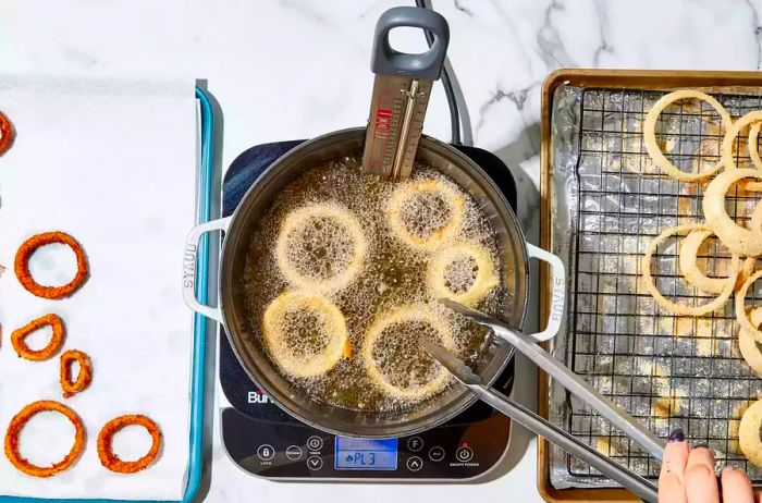 A large pot with a thermometer, frying onion rings in hot oil, next to a baking sheet lined with paper towels to drain the fried onion rings