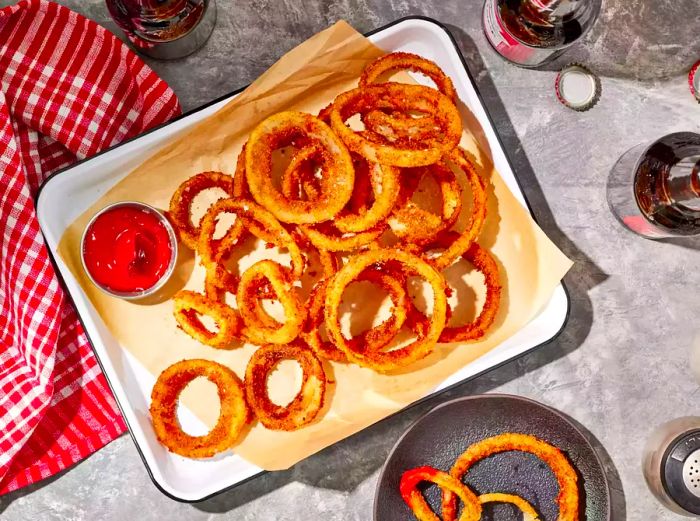 A parchment paper-lined tray filled with classic onion rings, served with a small bowl of ketchup and bottles of beer