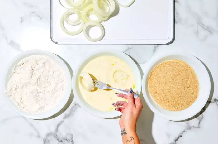 A hand holding a fork, dipping a flour-coated onion slice into a bowl of egg mixture at a breading station