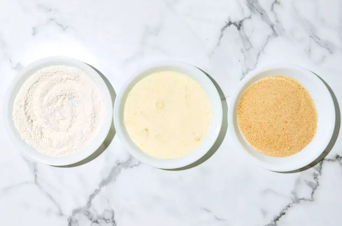 A breading station set up with three bowls: one with a flour mixture, another with an egg mixture, and the third with breadcrumbs