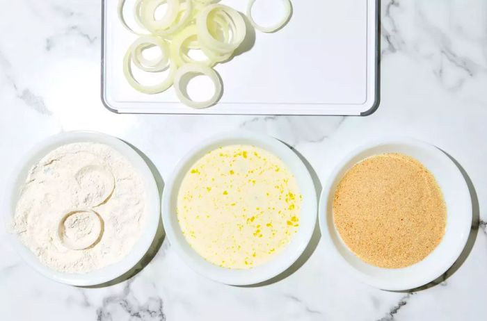 A cutting board with sliced onions at a breading station, with one bowl of flour and onion slices being dipped into it