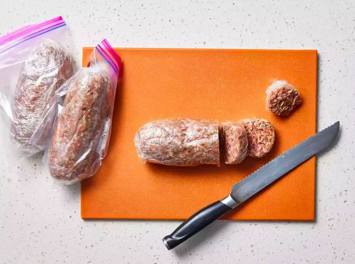 A cutting board with a log of seasoned sausage, wrapped in plastic, being sliced into patties