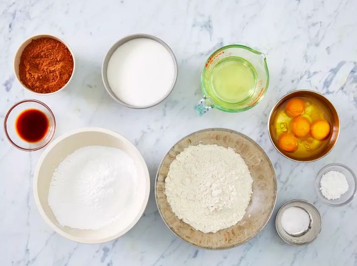 An overhead shot of the ingredients for chocolate crinkle cookies, arranged in small bowls on a marble kitchen countertop.