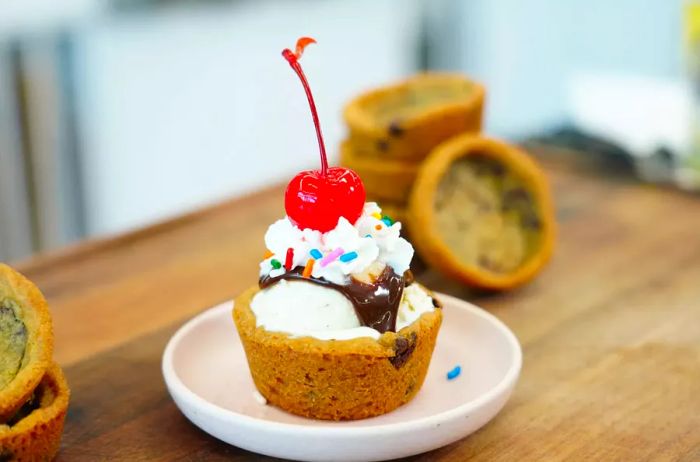 A chocolate chip cookie cup filled with vanilla ice cream, topped with chocolate syrup, whipped cream, candy sprinkles, and a maraschino cherry.