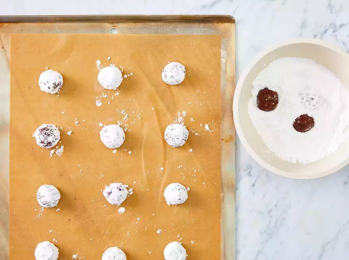 An overhead shot of chocolate crinkle cookie dough being rolled in powdered sugar and arranged on a prepared baking tray.