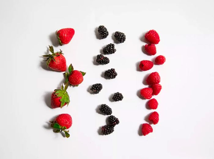 A variety of raspberries, strawberries, and blackberries arranged in neat lines on a clean white surface.