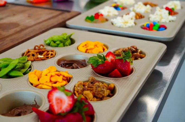 Muffin tins filled with a variety of snacks and dips: nuts, fruits, edamame, pretzels, candies, popcorn, and chocolate.