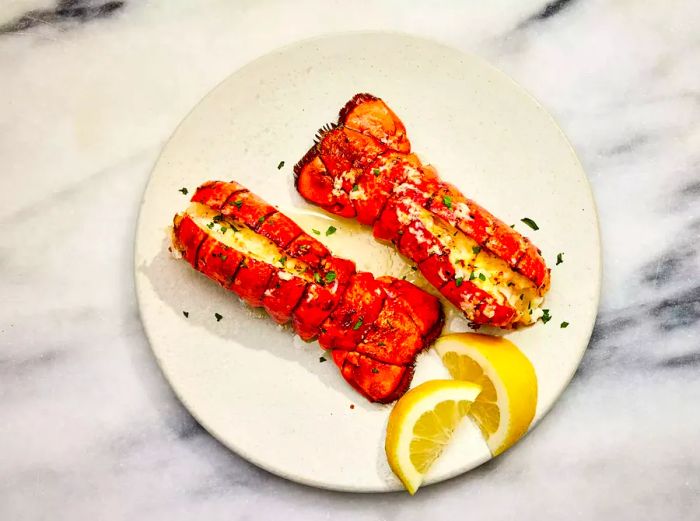 A plate featuring two lobster tails, topped with garlic-lemon butter and chopped parsley, served with wedges of lemon