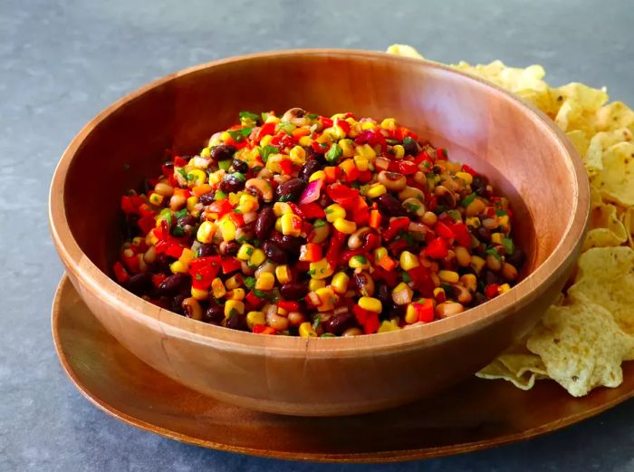 A wooden bowl of cowboy caviar served with a scoop of tortilla chips on a wooden platter.