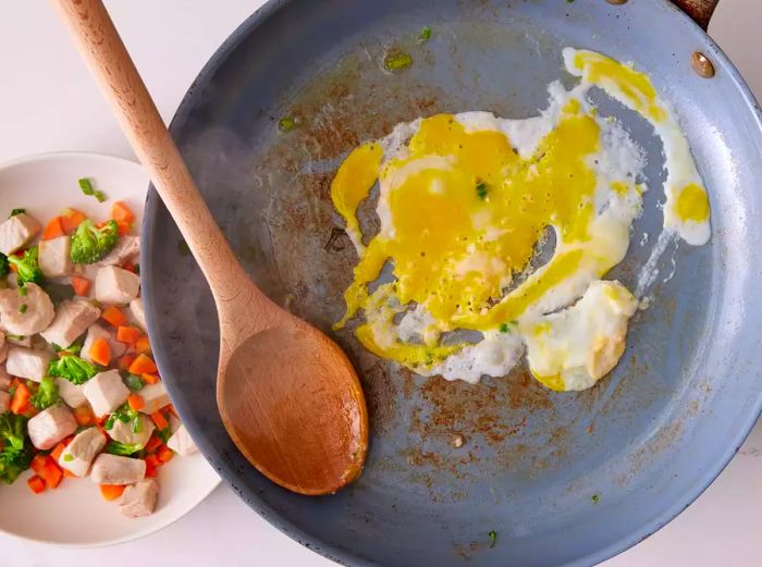 A large skillet with scrambled eggs, next to a bowl containing cooked pork, broccoli, carrots, and green onions.