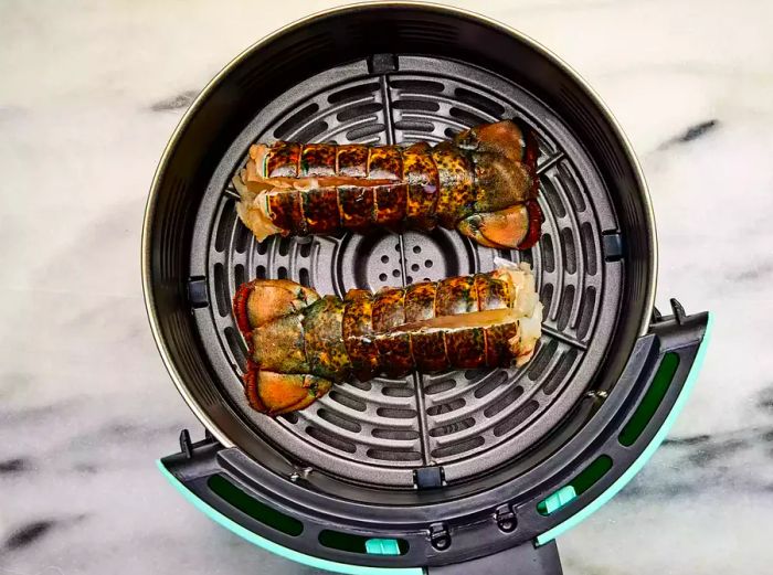 An air fryer basket holding two lobster tails with their shells cut open