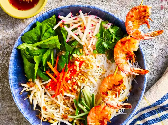 A top-down view of a vermicelli noodle bowl with shrimp skewers, vegetables, and peanuts.